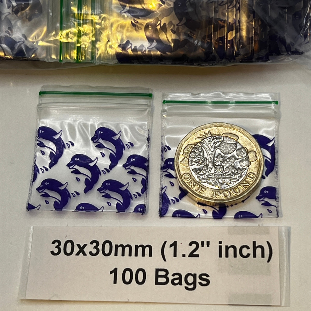 Two small resealable plastic bags with blue dolphin prints, one containing a gold coin for scale. A label below reads '30x30mm (1.2 inch)